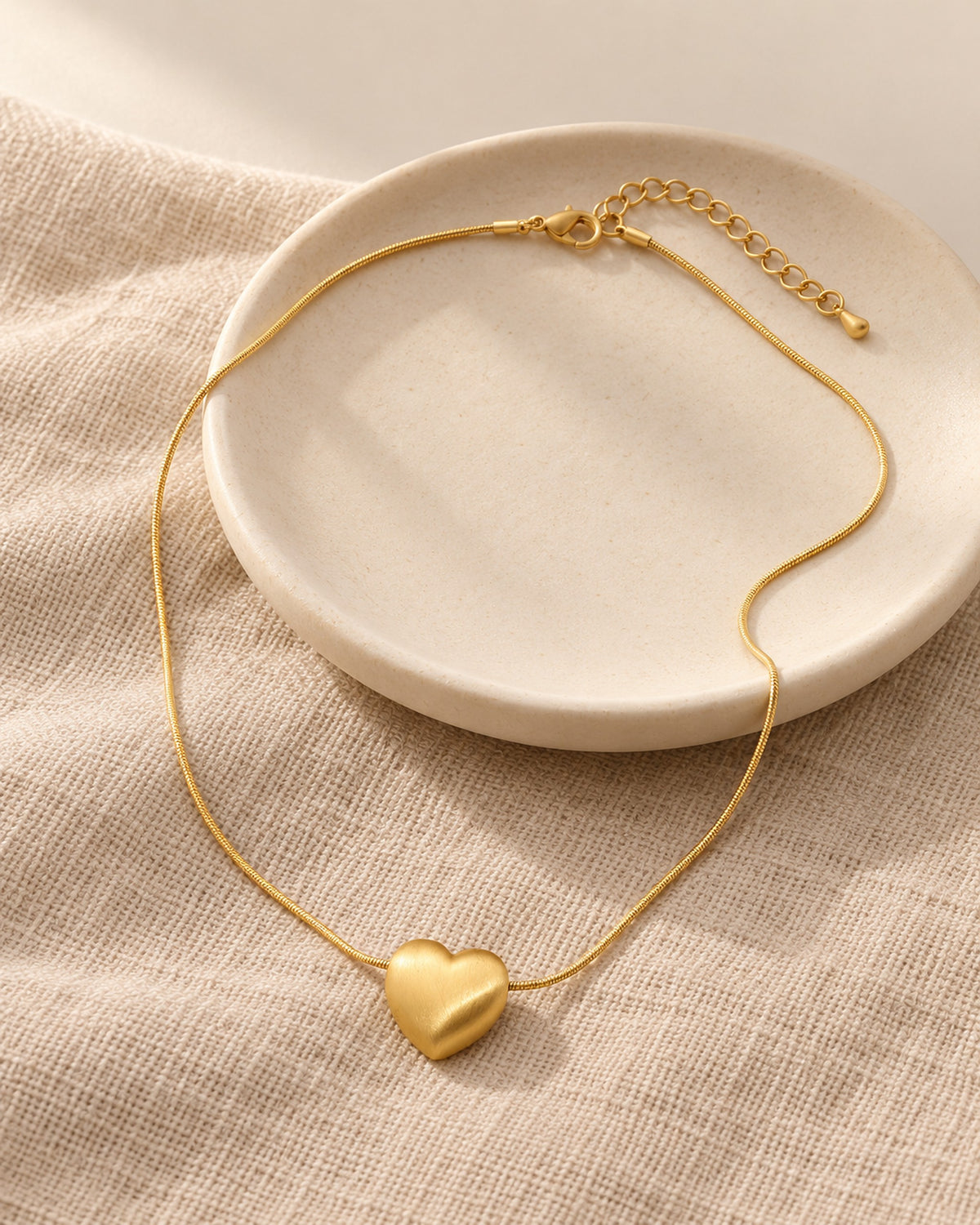 Héloïse brushed matte heart pendant necklace on snake chain in Gold, still life on linen